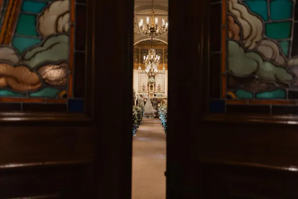A cerimônia vista pela porta entreaberta da igreja, entre vitrais, com os noivos no altar