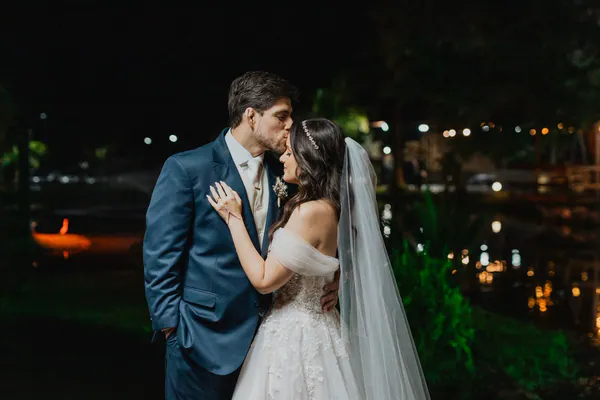 À noite, o noivo beija a testa da noiva, com luzes desfocadas ao fundo