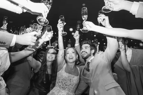 Em preto e branco, o brinde na festa, com os noivos rindo entre as taças