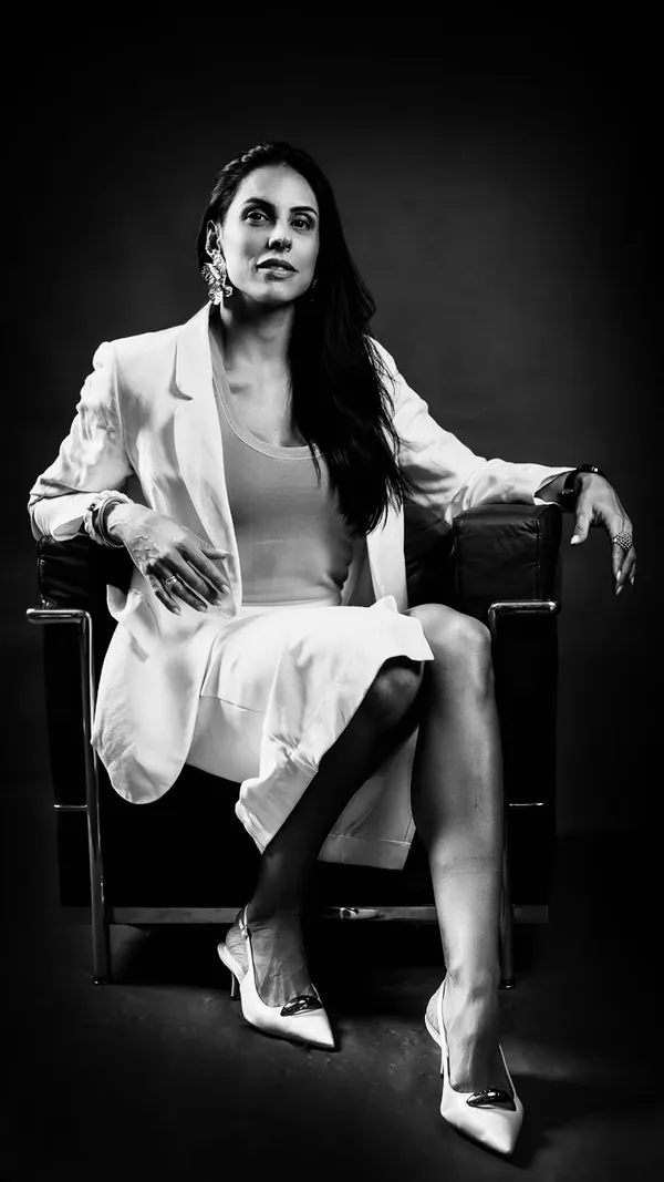 Em preto e branco, mulher de blazer branco sentada numa poltrona, de corpo inteiro
