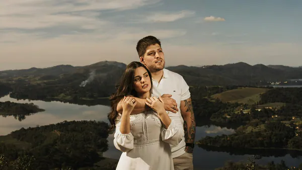 Ensaio de casal ao ar livre: ele a abraça por trás, no alto de um morro, com um lago e montanhas ao fundo