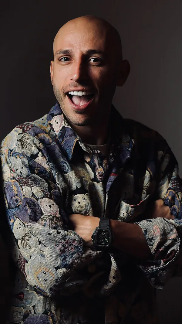 Homem rindo de braços cruzados, com camisa estampada, sobre fundo escuro